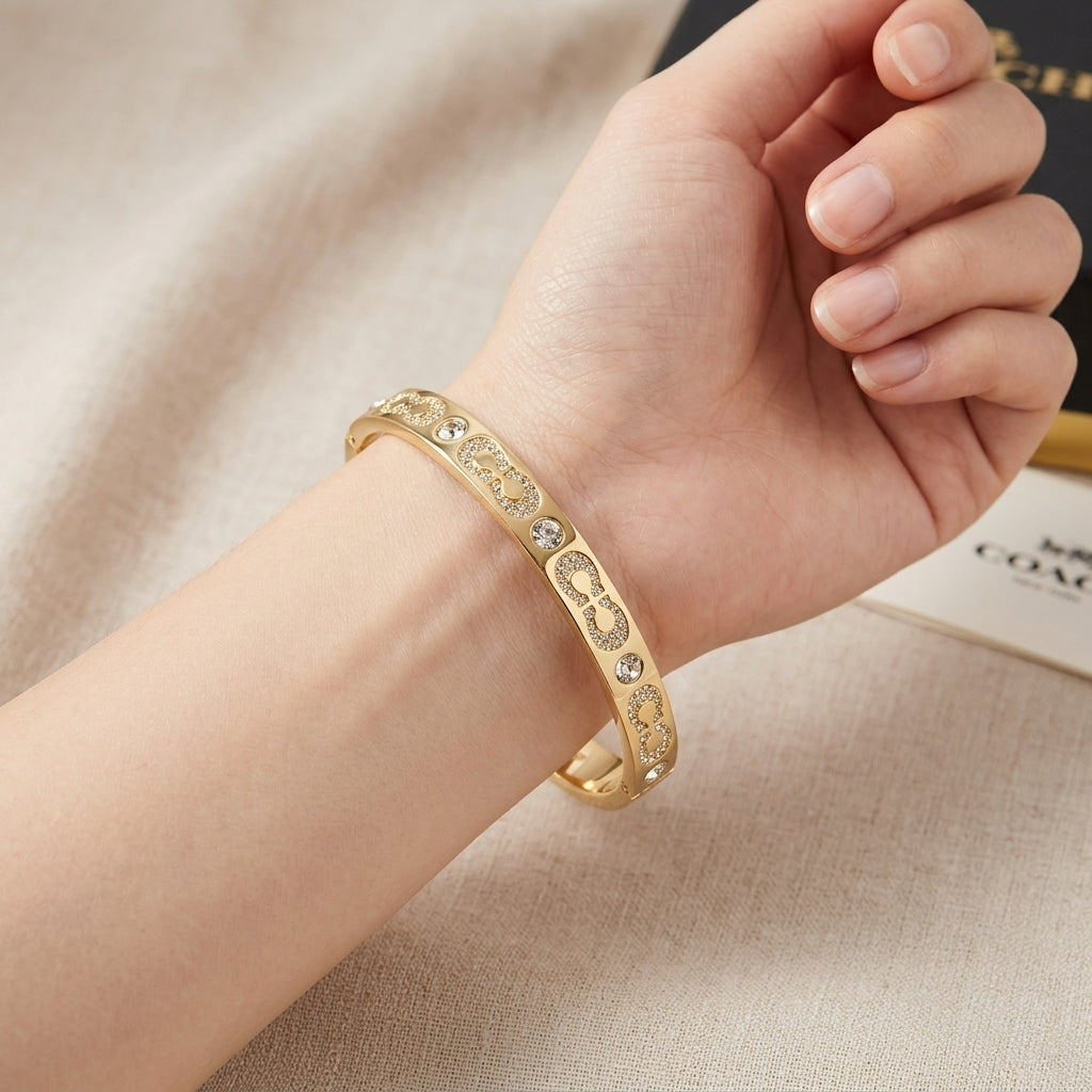 A gold-plated bangle bracelet with engraved floral patterns and small clear rhinestones, shown worn on a person's wrist against a beige fabric background.