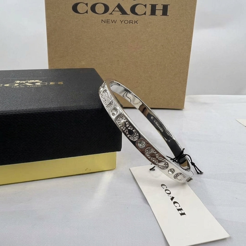 A silver-tone Coach bangle bracelet with engraved script and crystal-like details, displayed next to its black and gold presentation box and a white Coach tag, with a brown cardboard box in the background.