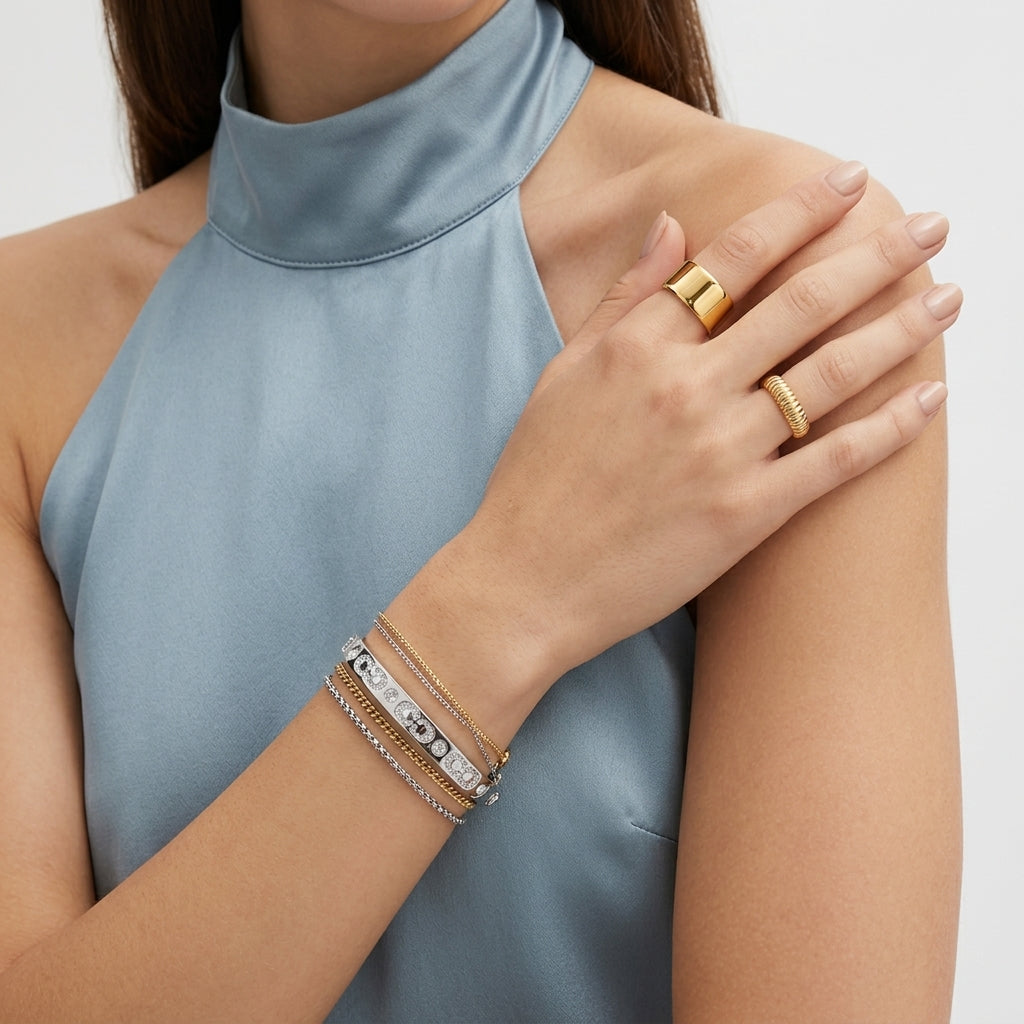 A close-up of a model wearing gold rings and stacked bracelets on her wrist, wearing a light blue satin halter top against a clean white background.