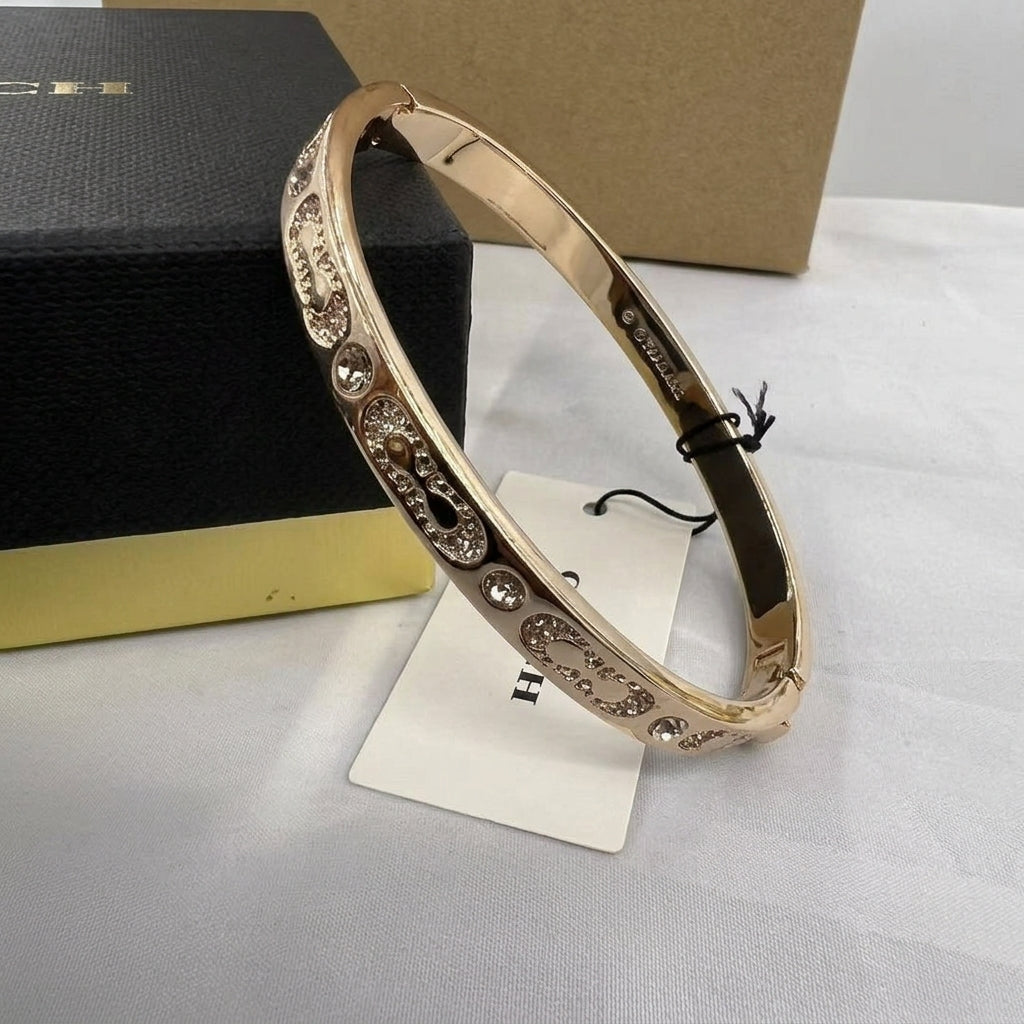 A rose gold bangle bracelet with clear rhinestone accents, displayed on a white stand with its black and gold H.E.