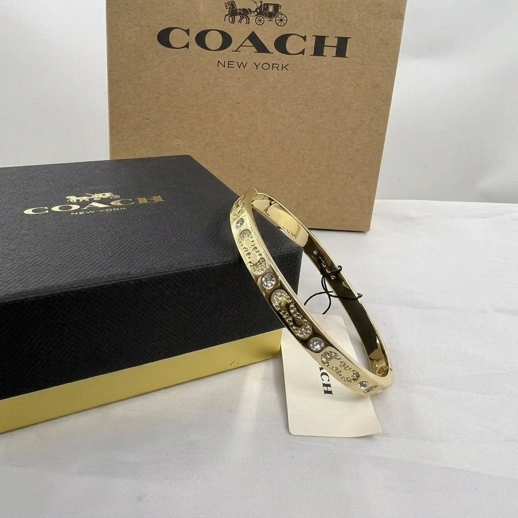 A gold-tone Coach bangle bracelet with clear crystal embellishments, displayed next to its black and gold branded box and a brown cardboard Coach box on a white surface.