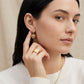 A close-up lifestyle shot of a woman wearing a small gold heart-shaped dangle earring and a wide gold ring, wearing a white ribbed sweater against a soft beige background.