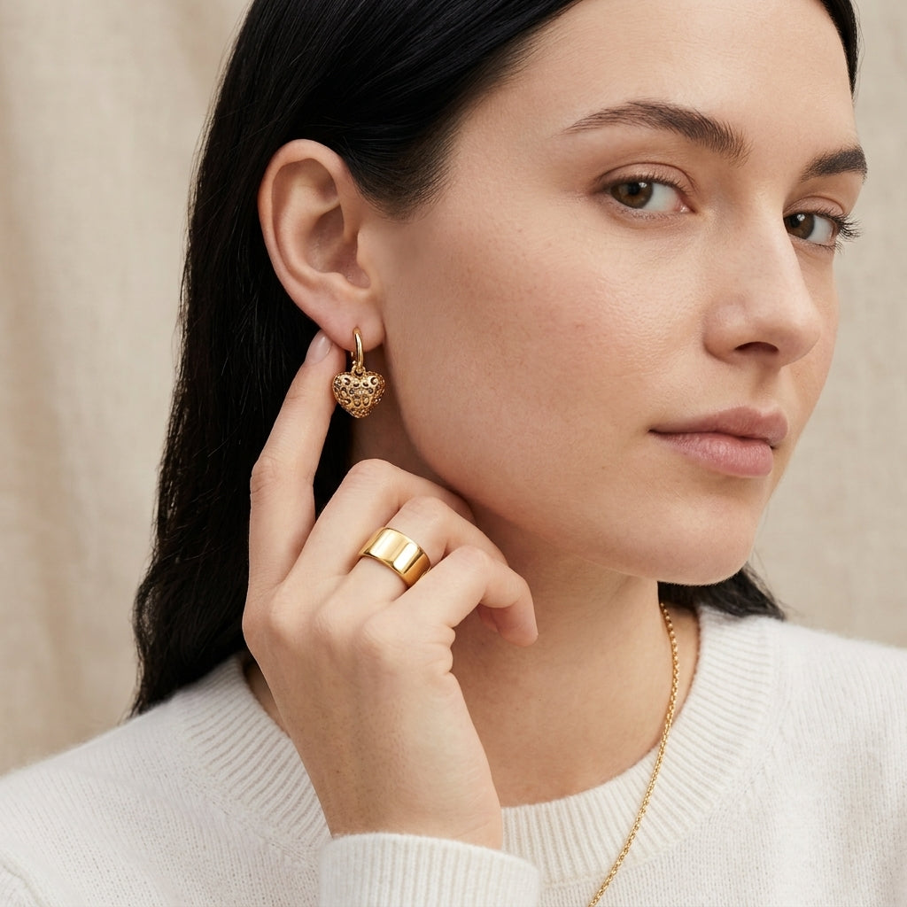 A close-up lifestyle shot of a woman wearing a small gold heart-shaped dangle earring and a wide gold ring, wearing a white ribbed sweater against a soft beige background.