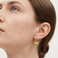 A close-up of a woman's ear wearing a small gold hoop earring with a heart-shaped charm covered in colorful enamel dots.