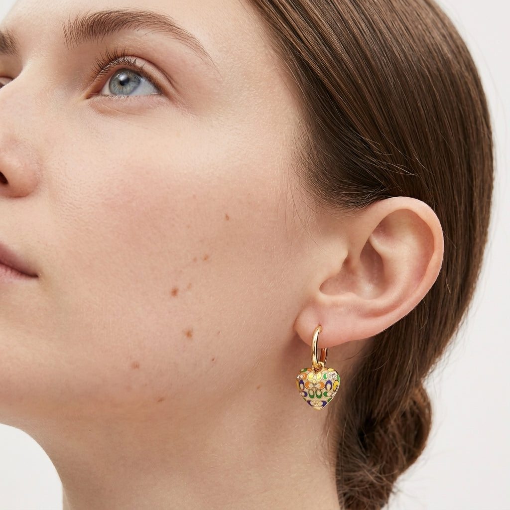 A close-up of a woman's ear wearing a small gold hoop earring with a heart-shaped charm covered in colorful enamel dots.