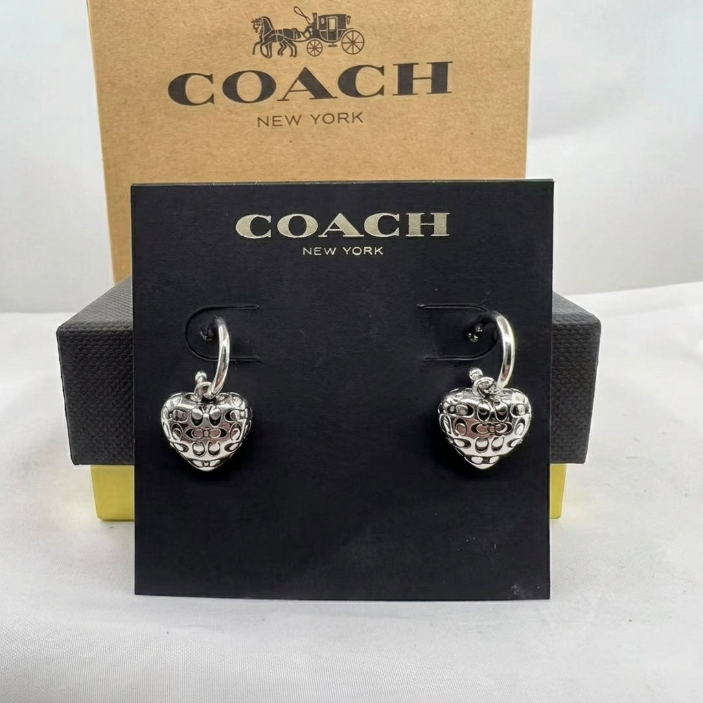 A pair of silver heart-shaped drop earrings displayed on a black Coach New York card, with the branded brown box behind it on a white surface.