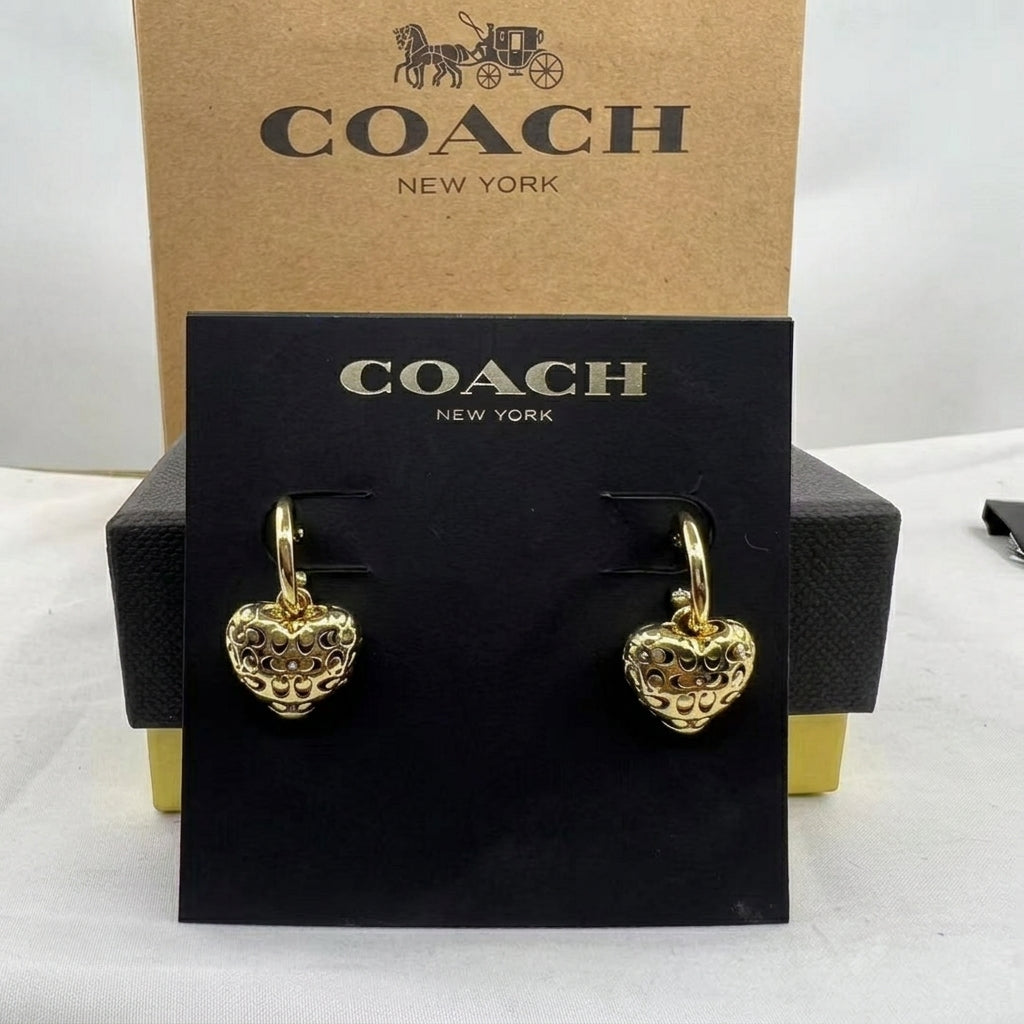 A pair of gold heart-shaped drop earrings with textured detailing, displayed on a black Coach New York card, with the brown Coach New York box behind it.