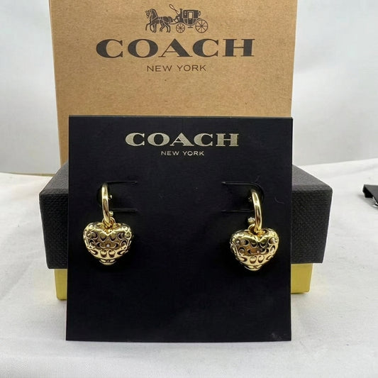 A pair of gold heart-shaped drop earrings with textured detailing, displayed on a black Coach New York card, with the brown Coach New York box behind it.
