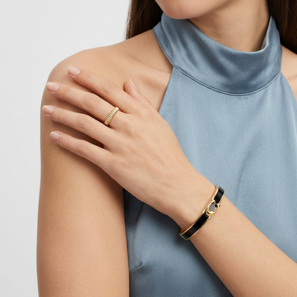 A close-up of a woman wearing a slim gold ring on her finger and a matching gold bangle bracelet with a black band on her wrist, wearing a light blue satin high-neck top against a light gray background.
