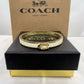 A Coach New York gold-tone and white bangle bracelet displayed on a black presentation box with a brown Coach box behind it.