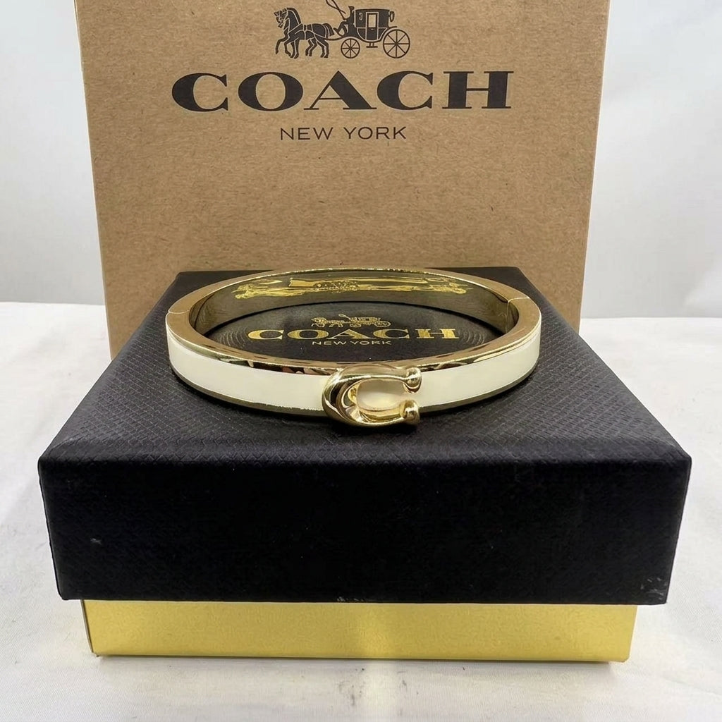 A Coach New York gold-tone and white bangle bracelet displayed on a black presentation box with a brown Coach box behind it.