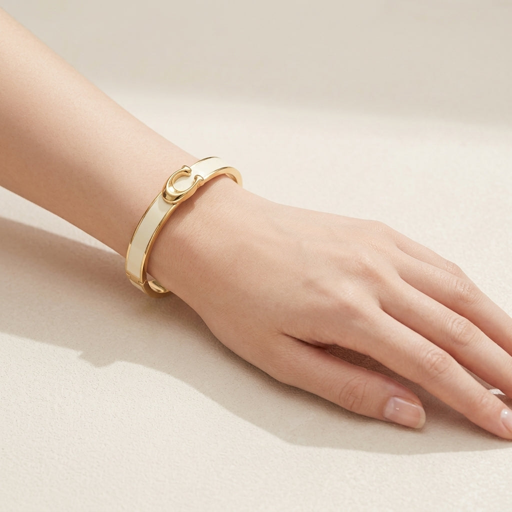 A gold bangle bracelet with a white enamel inlay and a small central gold charm, shown worn on a person's wrist against a neutral beige background.