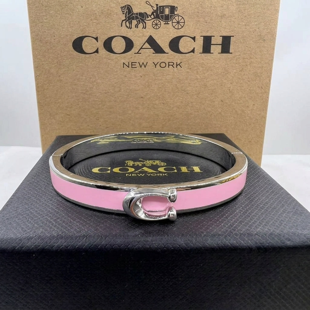 A pink and silver-tone Coach New York bangle bracelet displayed on a black presentation box, with the branded kraft box standing behind it.