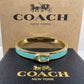 A Coach New York bangle bracelet with a gold-tone metal band, a turquoise blue enamel inlay, and the signature C logo clasp, displayed in front of its brown cardboard box and black presentation box.