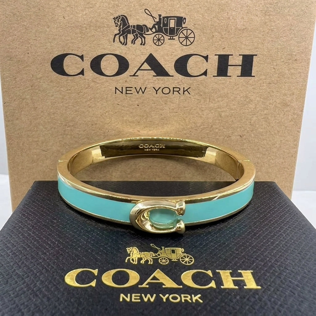A Coach New York bangle bracelet with a gold-tone metal band, a turquoise blue enamel inlay, and the signature C logo clasp, displayed in front of its brown cardboard box and black presentation box.