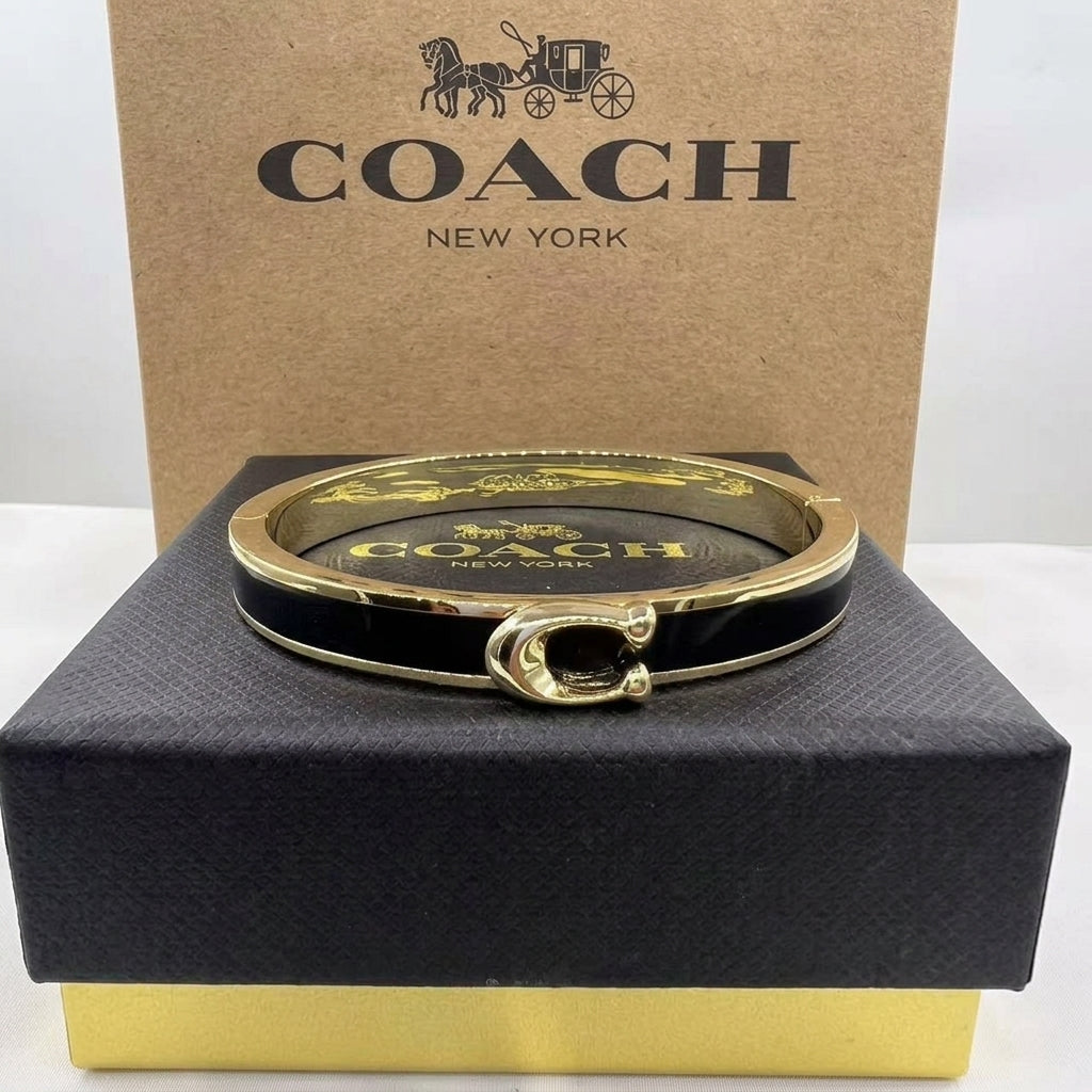 A Coach New York hinged bangle bracelet with a black enamel interior and gold-tone metal exterior, displayed on top of its black presentation box with the branded brown cardboard box behind it.
