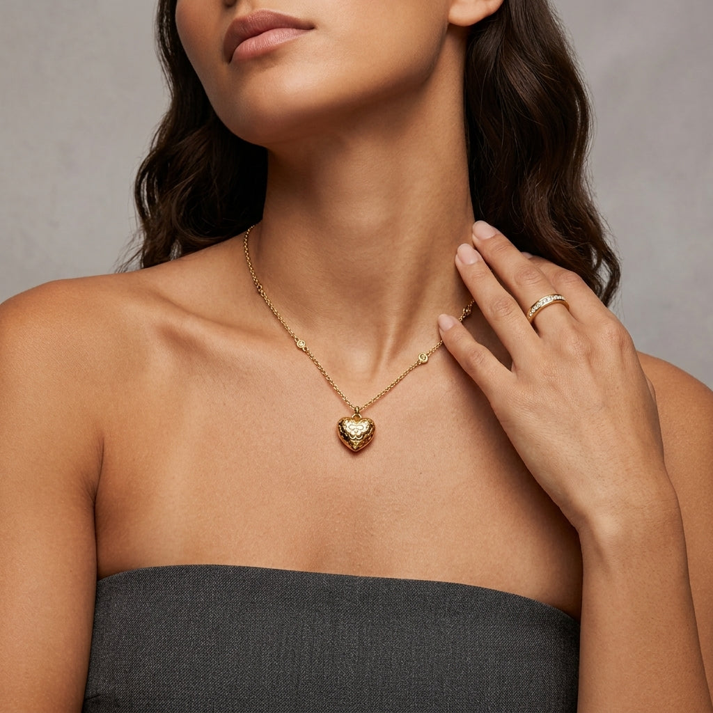 A close-up of a woman wearing a delicate gold chain necklace with a textured heart-shaped pendant, paired with a gold ring on her finger, set against a neutral gray background.