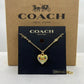 A gold-tone Coach necklace with a colorful enamel heart pendant, displayed on a black Coach card in front of a brown Coach New York box.