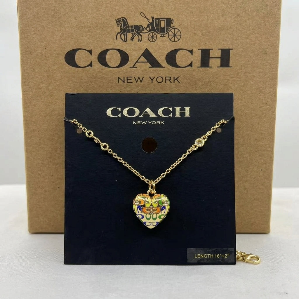 A gold-tone Coach necklace with a colorful enamel heart pendant, displayed on a black Coach card in front of a brown Coach New York box.