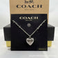 A silver heart-shaped pendant necklace with a floral filigree design, displayed on a black Coach New York card, with a brown Coach New York box behind it.