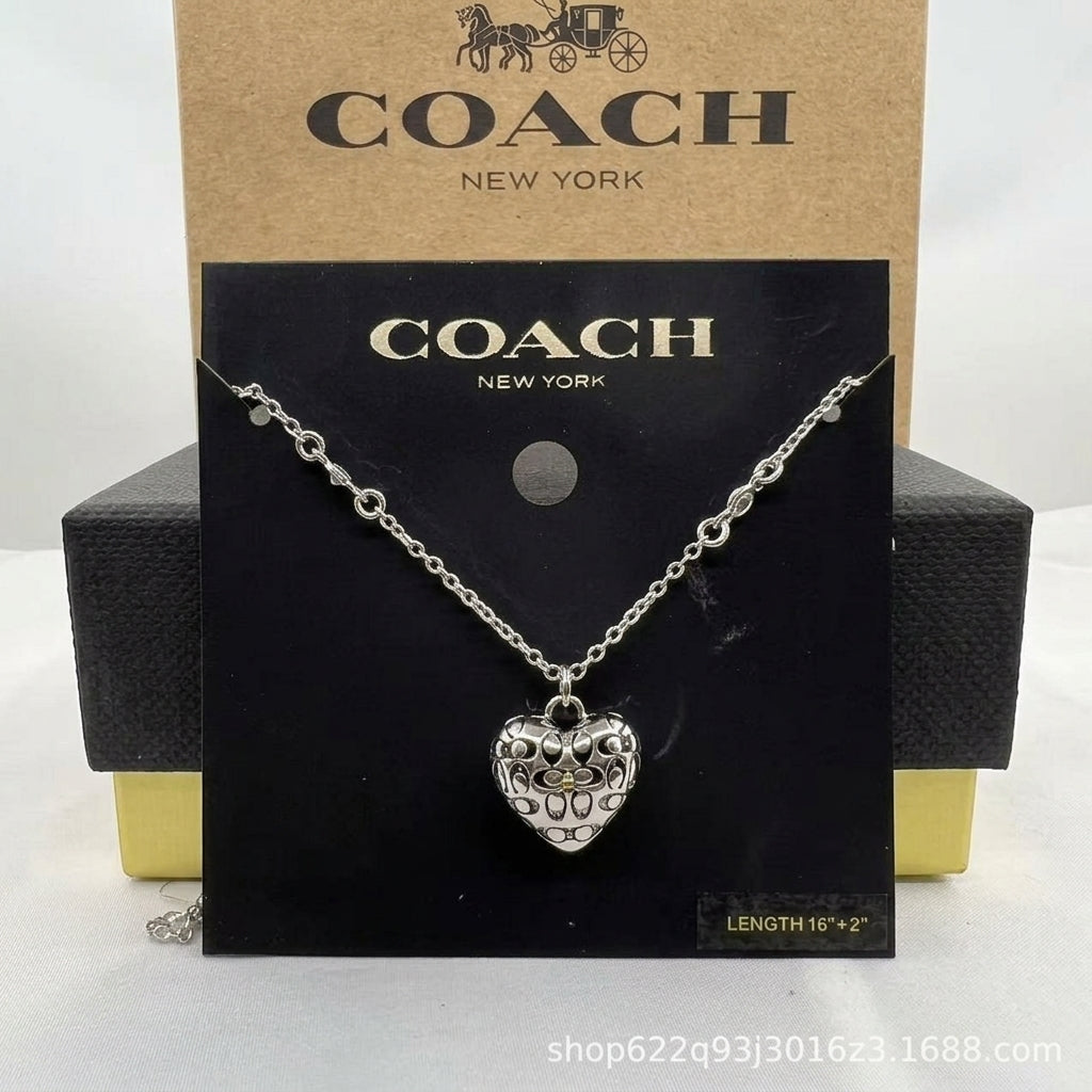 A silver heart-shaped pendant necklace with a floral filigree design, displayed on a black Coach New York card, with a brown Coach New York box behind it.
