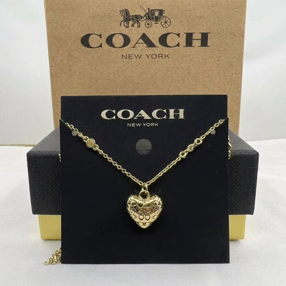 A gold-tone Coach necklace with a heart-shaped pendant displayed on a black Coach card, with the Coach New York box behind it.