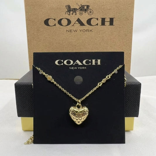 A gold-tone Coach necklace with a heart-shaped pendant displayed on a black Coach card, with the Coach New York box behind it.