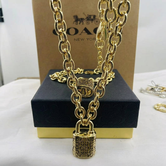 A gold-tone Coach necklace with a padlock pendant, displayed on a black presentation box with a brown Coach box behind it.