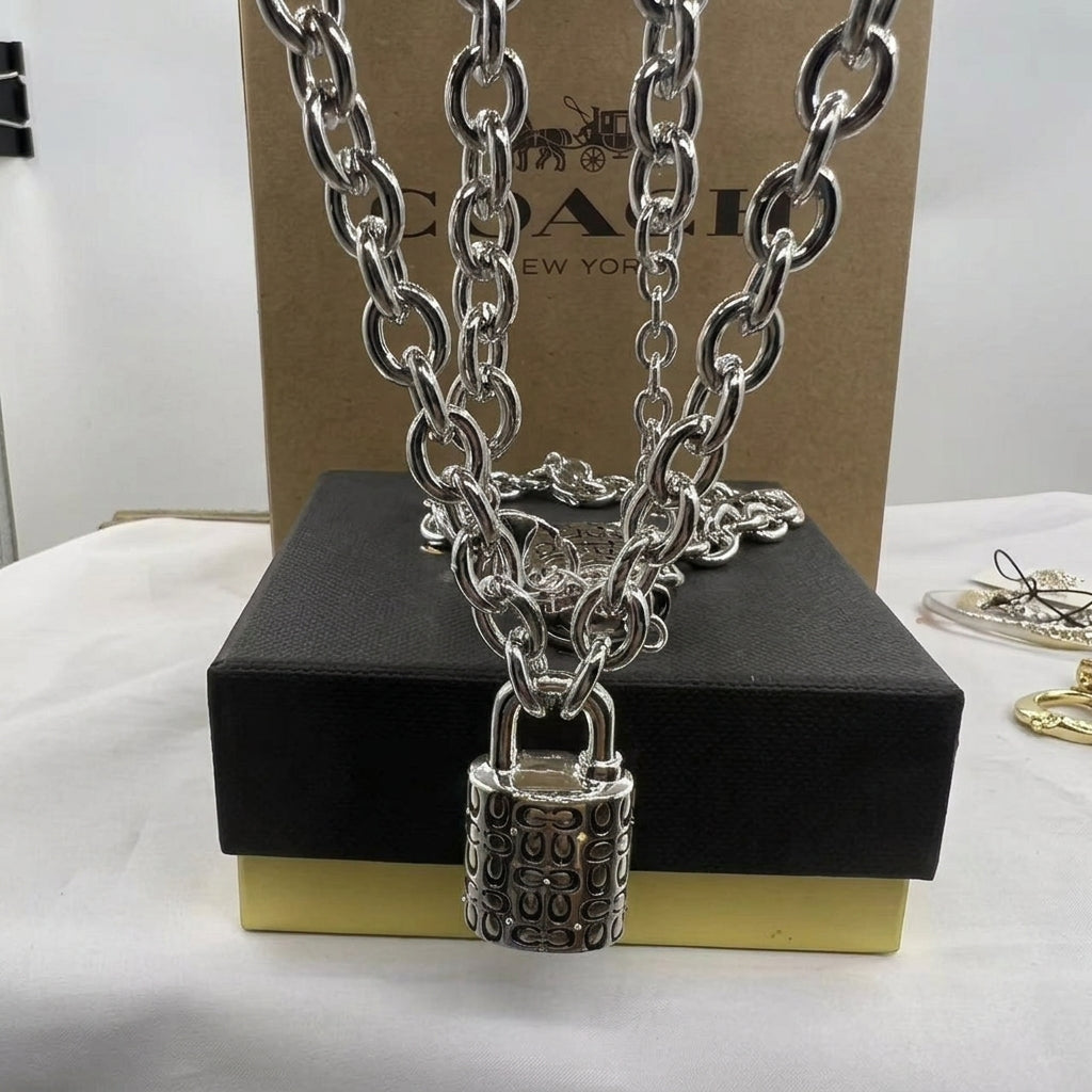 A silver-tone padlock necklace with interlocking 'CO' initials on the lock, draped over a black and gold Coach gift box with a Coach box in the background.