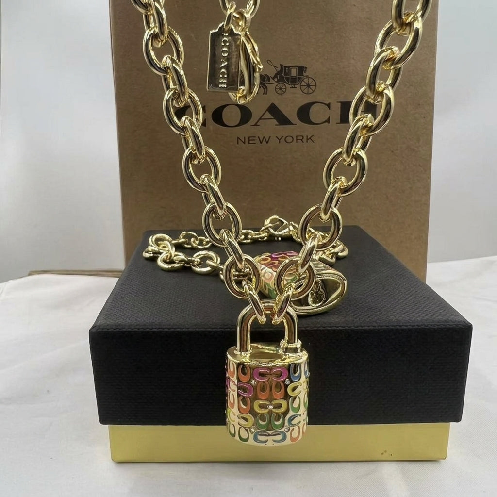 A gold-tone Coach chain necklace with a padlock pendant, displayed on a black presentation box with a Coach New York box behind it.