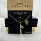 A gold-tone Snoopy and Woodstock necklace displayed on a black Coach New York card, with the Coach box behind it on a light surface.