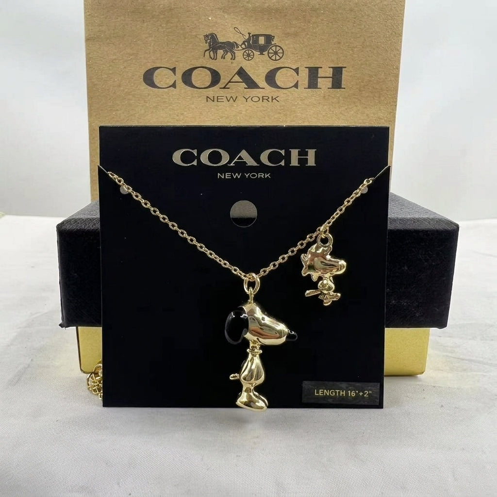 A gold-tone Snoopy and Woodstock necklace displayed on a black Coach New York card, with the Coach box behind it on a light surface.