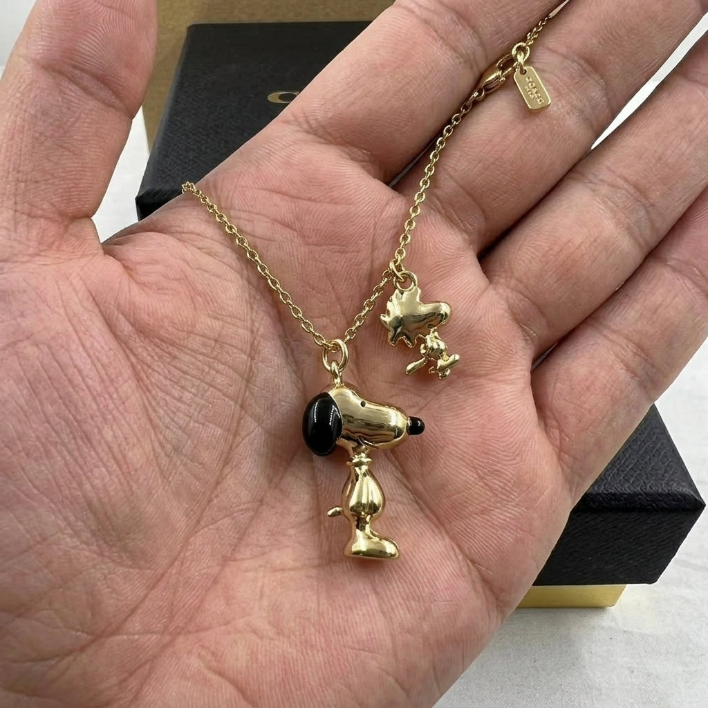 A gold-tone necklace with two character pendants, one with a black nose and one with a black ear, displayed on an open hand with a black jewelry box in the background.