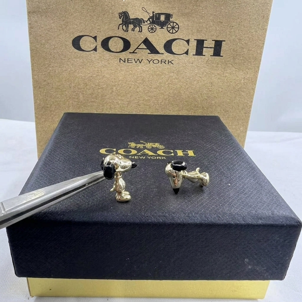 Gold-tone Snoopy and Charlie Brown cufflinks displayed on a black Coach New York box, with tweezers holding one cufflink in front of a Coach New York branded box.