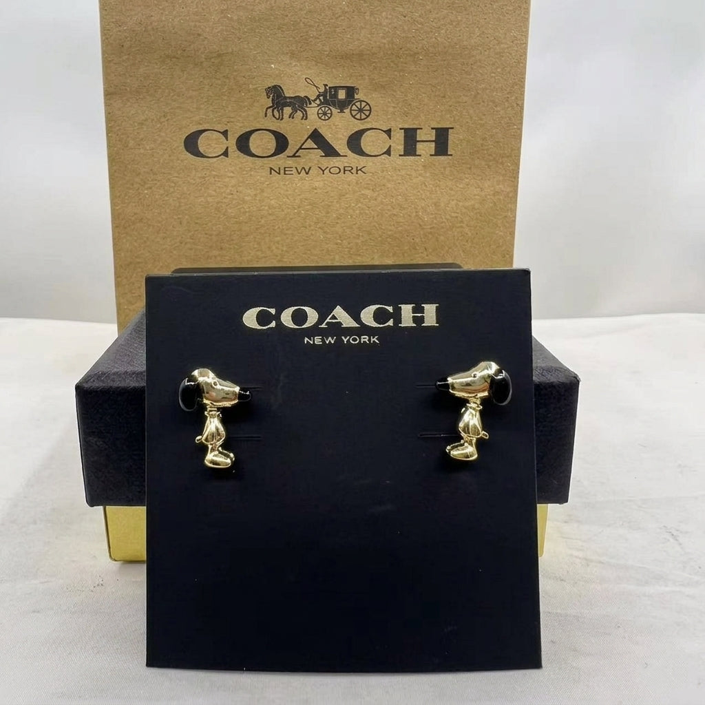 A pair of gold-tone dog stud earrings displayed on a black Coach New York card, with the black and kraft Coach packaging behind it on a white surface.