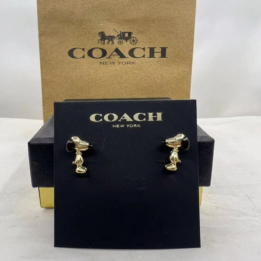 A pair of gold-tone dog stud earrings displayed on a black Coach New York card, with the black and kraft Coach packaging behind it on a white surface.