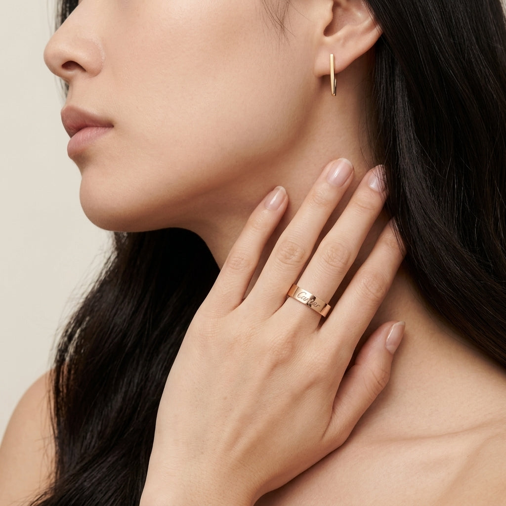 A close-up lifestyle shot of a woman wearing a slim gold band ring with a small central gemstone on her ring finger, alongside a matching gold hoop earring.