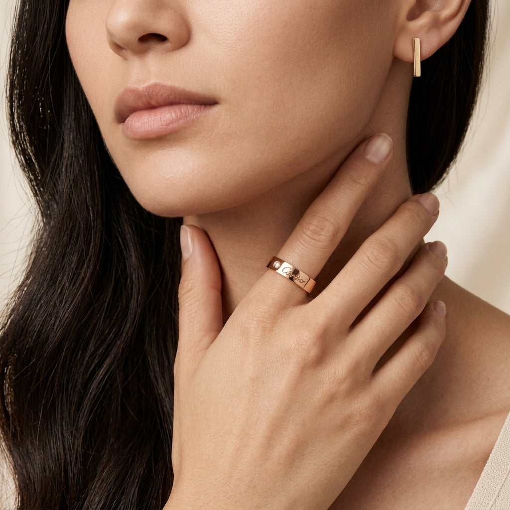 A close-up lifestyle shot of a model wearing a slim rose gold band ring on her finger, with her hand resting on her neck. She is also wearing a matching rose gold bar earring.