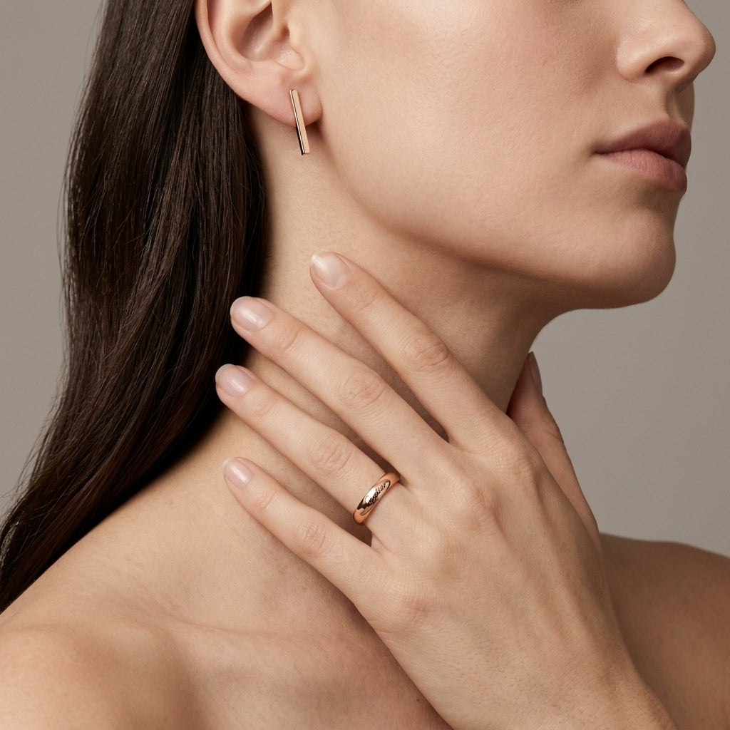 A close-up lifestyle shot of a model wearing a slim, polished rose gold band ring on her finger, with her hand resting on her neck. She also wears a matching rose gold bar stud earring.