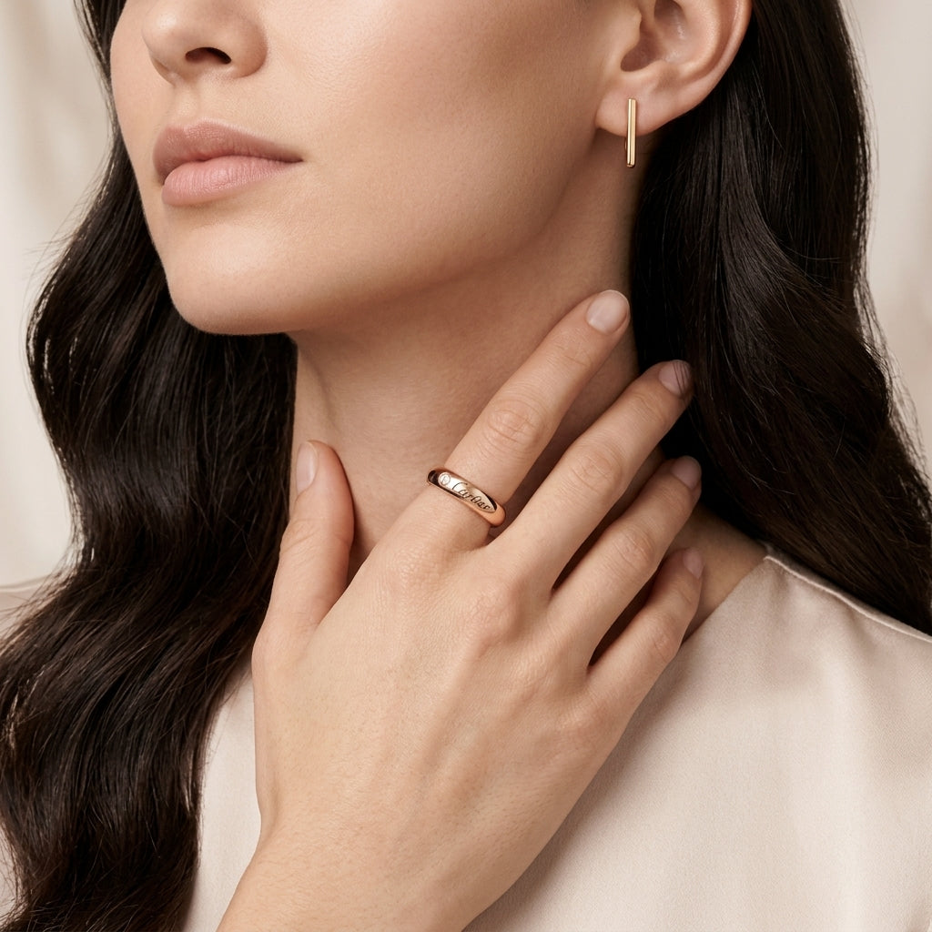 A close-up lifestyle shot of a woman wearing a slim rose gold band ring on her finger and a matching vertical bar earring in her ear, set against a soft beige background.