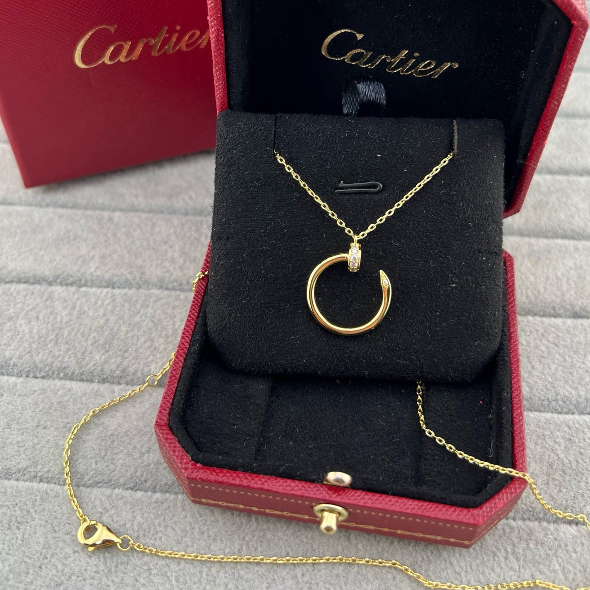 A gold Cartier necklace with a circular pendant displayed inside an open red Cartier jewelry box on a gray textured surface.