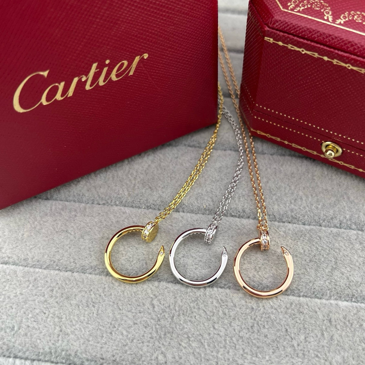 Three Cartier huggie hoop necklaces in gold, silver, and rose gold, displayed on a gray textured surface with red Cartier boxes in the background.
