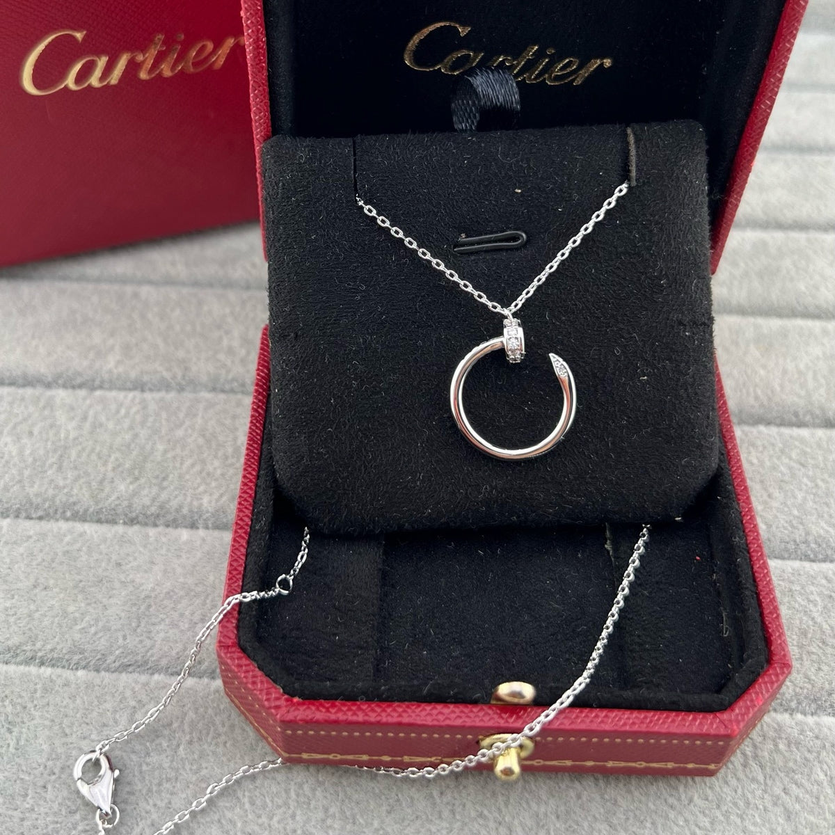 A Cartier necklace displayed inside an open red Cartier gift box on a black velvet cushion, with the necklace featuring a silver-tone circular pendant and a delicate chain with a small gold bead accent.