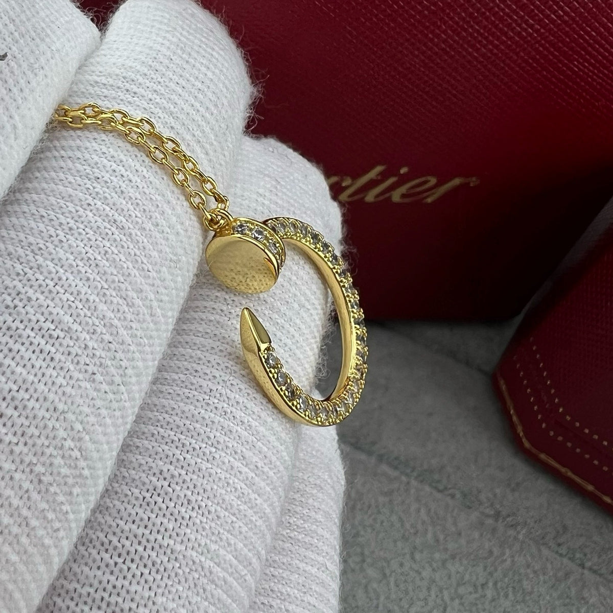 A gold Cartier bracelet with a circular pavé ring and a small heart charm, displayed on a white textured glove with a red Cartier box in the background.