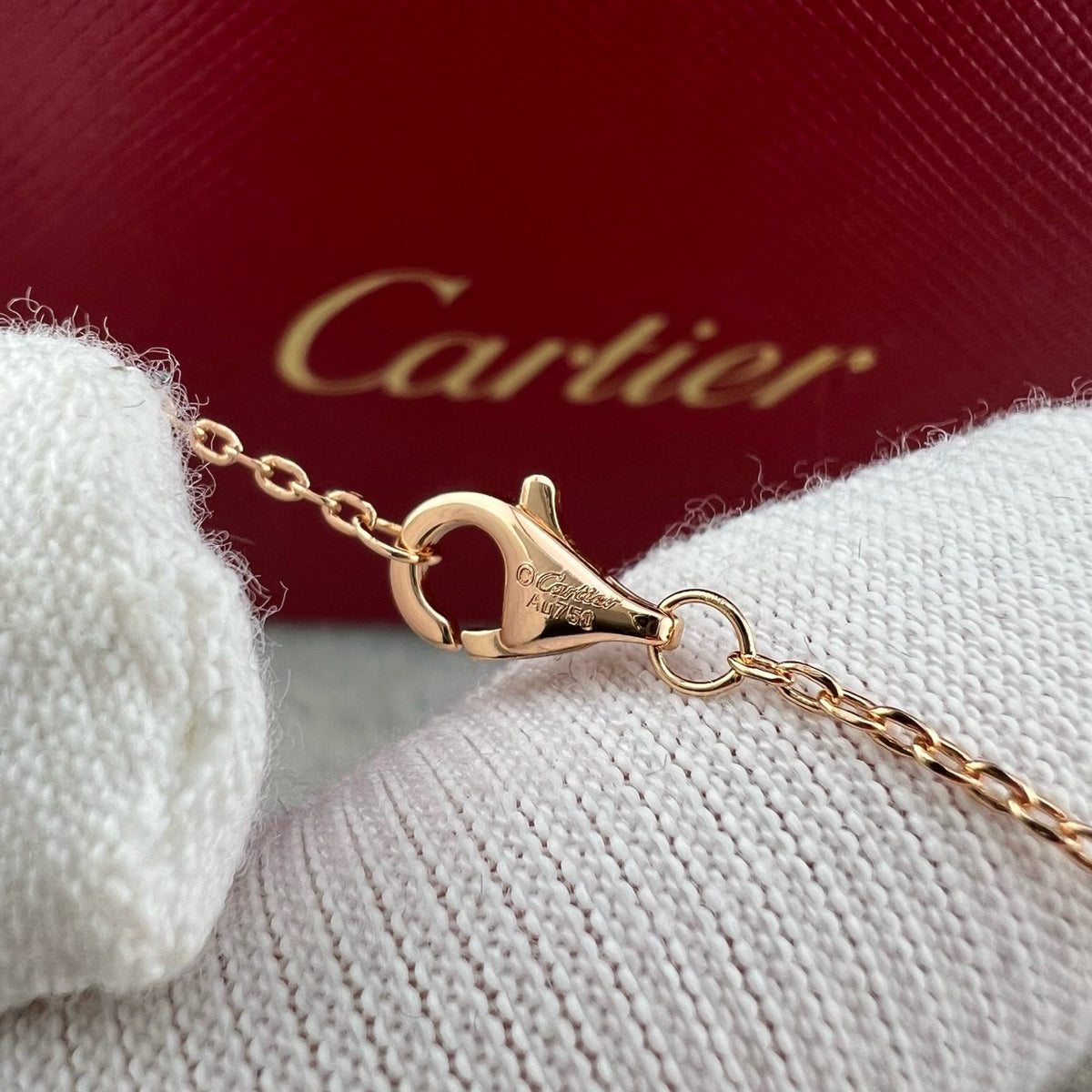 A close-up of a gold-tone Cartier chain with a lobster clasp resting on a white knit surface, with a red Cartier box blurred in the background.