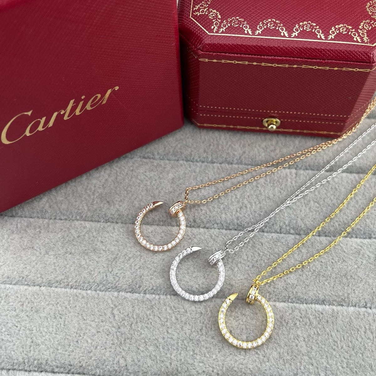 Three Cartier hoop necklaces with pavé-set round stones in gold, silver, and rose gold, displayed on a gray textured surface with red Cartier gift boxes in the background.