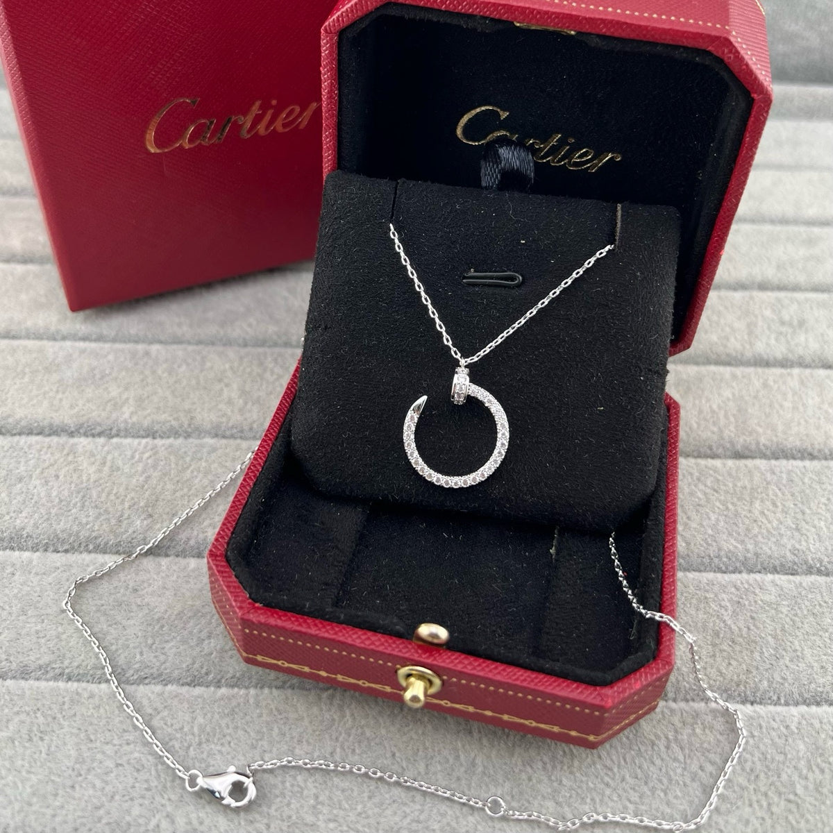 A silver crescent moon pendant necklace with a pavé-set top, displayed inside an open red Cartier jewelry box on a gray textured surface, with the Cartier box lid in the background.