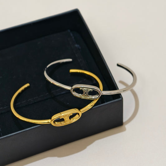 Two slim cuff bracelets with oval interlocking links, one in yellow gold and one in silver, displayed on a black jewelry box against a light beige background.