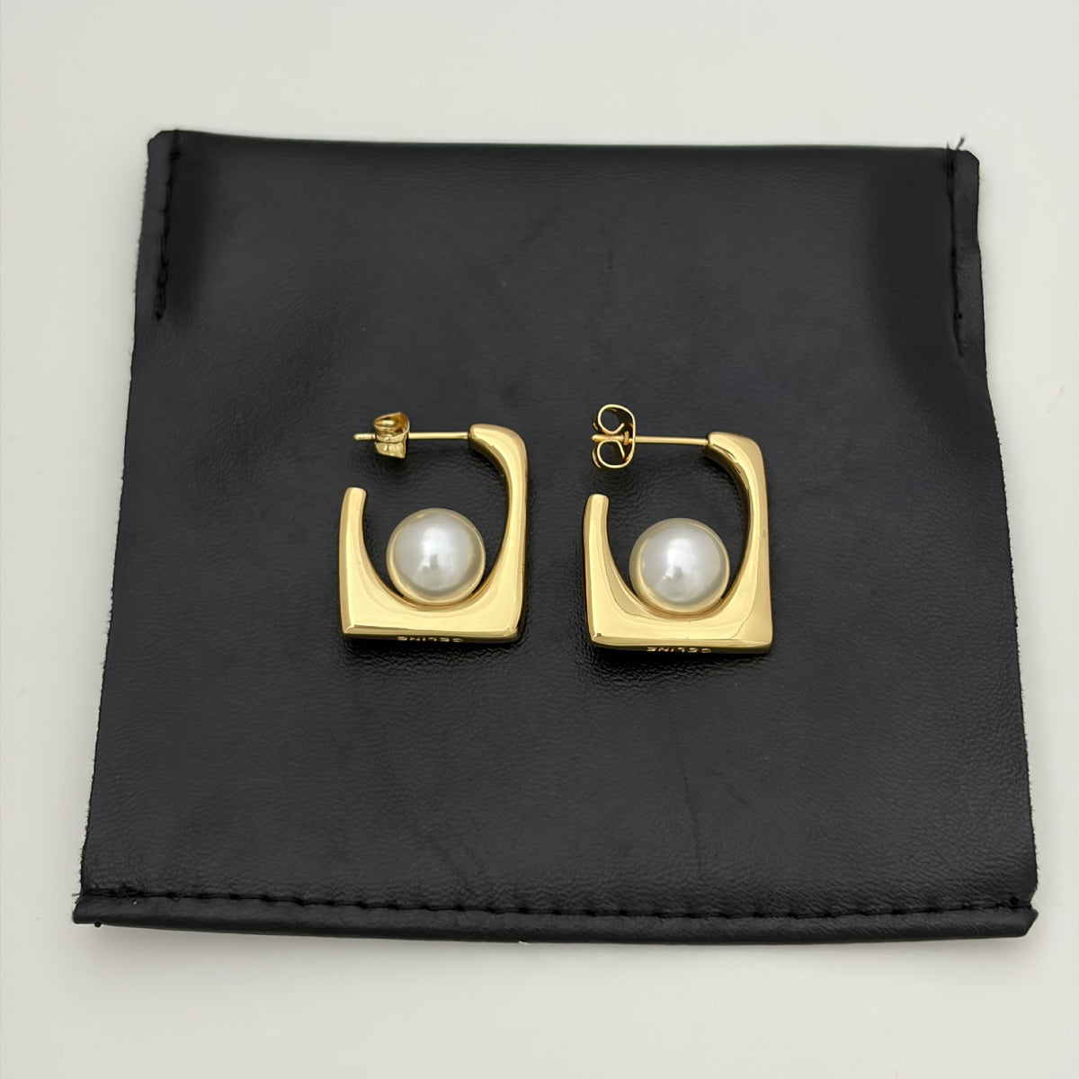 A pair of gold square hoop earrings with a central white pearl, displayed on a black textured fabric pouch against a light gray background.