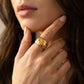 A close-up lifestyle shot of a woman wearing a delicate gold necklace with a circular interlocking logo pendant, along with a matching gold hoop earring and a simple gold band ring. She is wearing a cream-colored spaghetti strap top against a neutral light gray background.